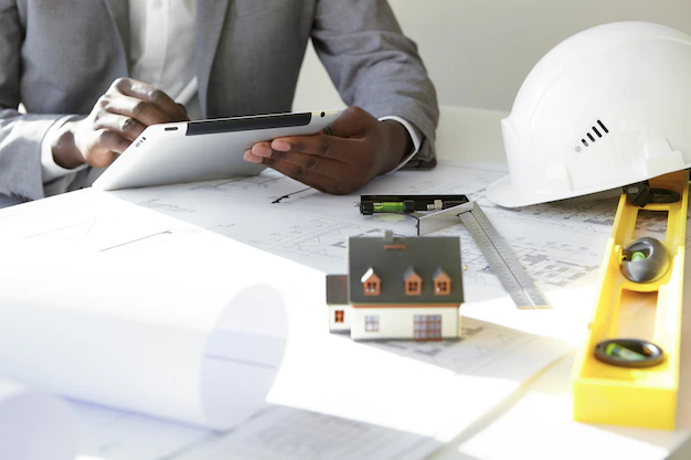 cropped-shot-dark-skinned-contractor-holding-touch-pad-entering-data-while-working-new-housing-project-sitting-desk-with-drawings-scale-model-house-blueprint-rolls-ruler-helmets_273609-5523